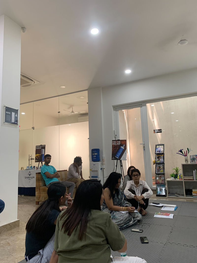 A casual group session inside a bright room with people sitting on the floor, surrounded by Savikalpa banners and products.4