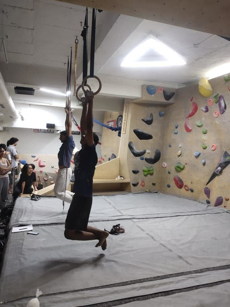 A man hanging from gymnastic rings in a climbing gym with climbing walls in the background at Boulder Blast by Savikalpa