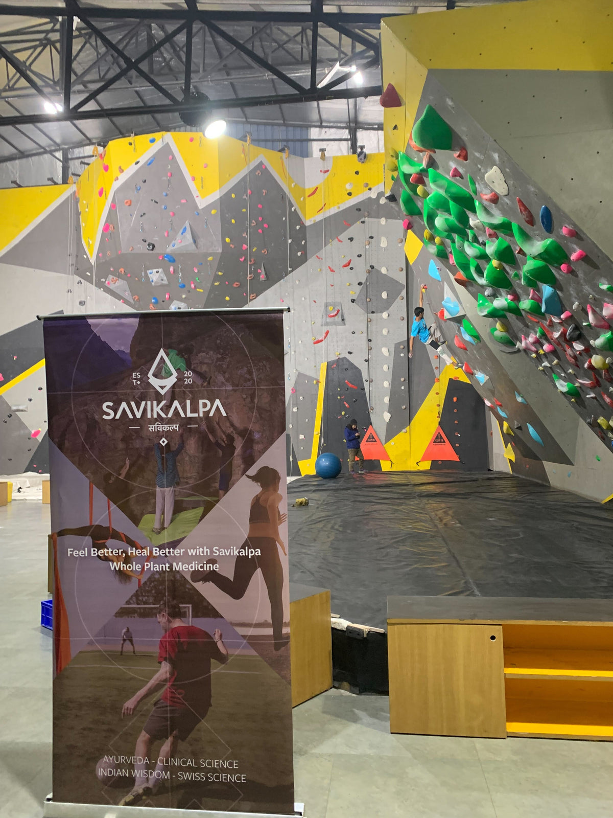 Indoor rock climbing facility, featuring promotional banner for Savikalpa focusing on wellness and plant-based medicine.